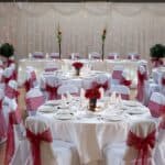 Tables and chairs with white cloths, red bows and red flowers set up with cutlery for an event