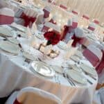 Table with red flowers in the middle, set up with cutlery and glasses. White chairs around the tables with red ribbon bows.